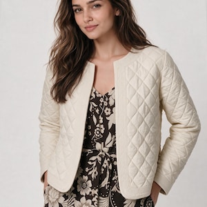 May include: A cream-colored quilted jacket with a diamond pattern and a round neckline, worn over a dark brown dress featuring a light-colored floral print. The jacket has long sleeves and a structured border along the front opening.