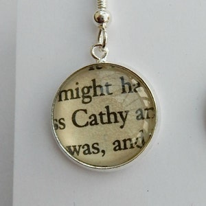 May include: Silver dangle earrings with a round glass charm featuring a printed text excerpt from a book. The text reads "might he Cathy and was, and".