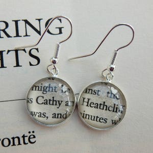 May include: Silver earrings with round glass charms featuring text from the book "Wuthering Heights" by Emily Bront&euml;. The text on the charms reads "might L ss Cathy and was, and" and "inst the Heathclif inutes W".