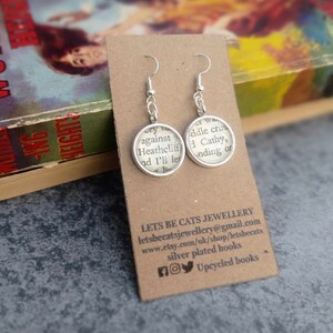 May include: A pair of silver earrings with round glass charms featuring text from a book. The text on the left charm reads "cry to against Heathcliff and I'll let it." The text on the right charm reads "of was middle crus Cathy, ending or".