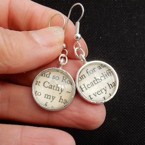 May include: Silver earrings with round glass charms featuring text from the novel "Wuthering Heights" by Emily Bront&euml;. The text on the left charm reads "and so Ro at Cathy - to my ha", and the text on the right charm reads "on for as Heathcliff t very ha".
