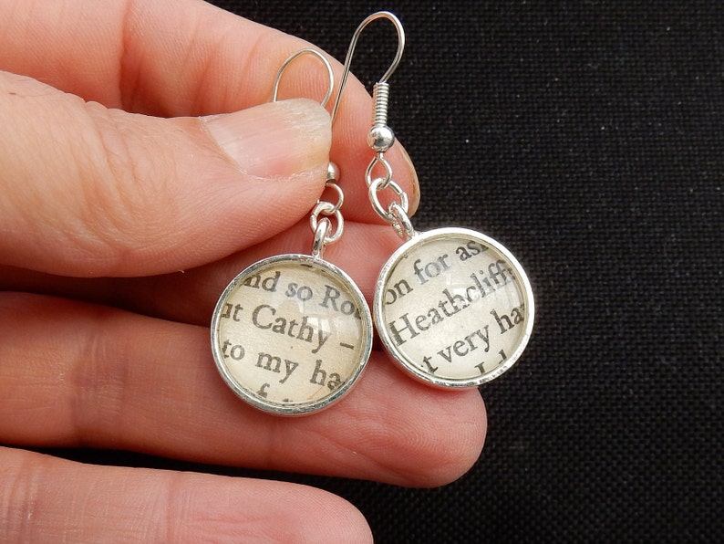 May include: Silver earrings with round glass charms featuring text from the novel "Wuthering Heights" by Emily Bront&euml;. The text on the left charm reads "and so Ro at Cathy - to my ha", and the text on the right charm reads "on for as Heathcliff t very ha".