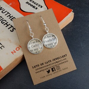 May include: A pair of silver dangle earrings with glass charms featuring text from a book. The text reads "and so Ro at Cathy to my la very ha". The earrings are displayed on a brown card with the text "LETS BE CATS JEWELLERY" and contact information.