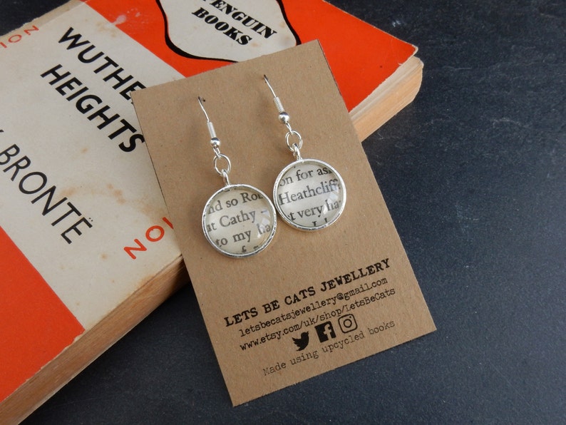 May include: A pair of silver dangle earrings with glass charms featuring text from a book. The text reads "and so Ro at Cathy to my la very ha". The earrings are displayed on a brown card with the text "LETS BE CATS JEWELLERY" and contact information.