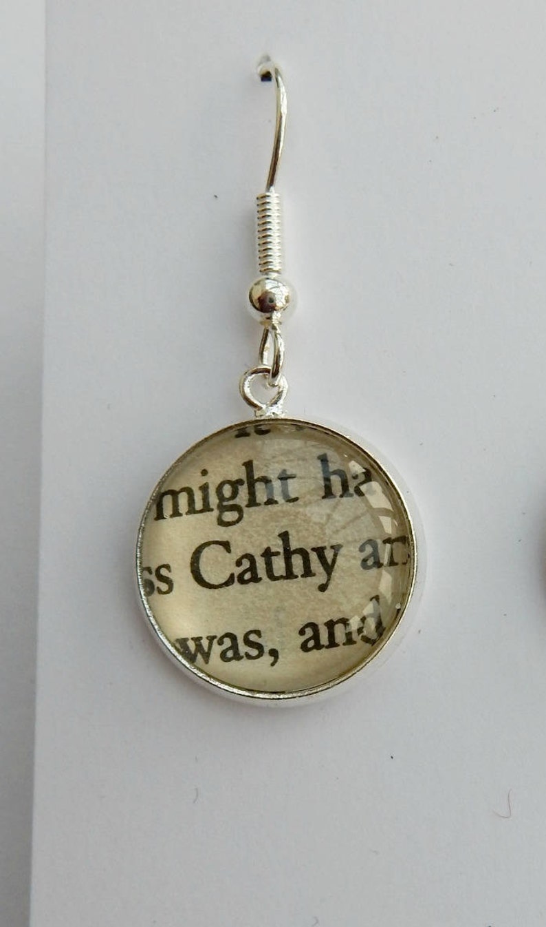 May include: Silver dangle earrings with a round glass charm featuring a printed text excerpt from a book. The text reads "might he Cathy and was, and".