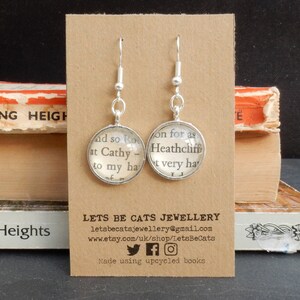 May include: Silver earrings with glass charms featuring text from the book "Wuthering Heights". The text on the charms reads "and so Ro at Cathy - to my ha" and "on for as Heathcliff t very ha". The earrings are displayed on a brown card with the text "LETS BE CATS JEWELLERY" and contact information.