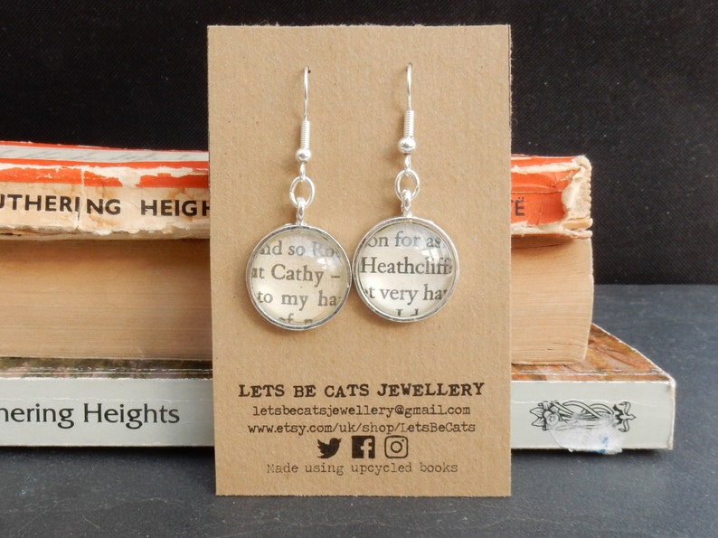 May include: Silver earrings with glass charms featuring text from the book "Wuthering Heights". The text on the charms reads "and so Ro at Cathy - to my ha" and "on for as Heathcliff t very ha". The earrings are displayed on a brown card with the text "LETS BE CATS JEWELLERY" and contact information.