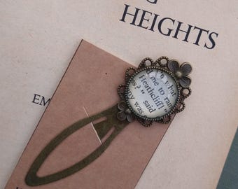 May include: A bronze bookmark with a round glass charm featuring a quote from the book "Wuthering Heights" by Emily Brontë. The quote reads: "Do what you like, I'm not afraid of you, Heathcliff!" The bookmark is on a brown background.