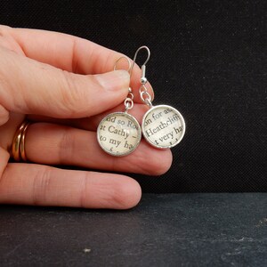 May include: Silver earrings with round glass charms featuring text from the book "Wuthering Heights" by Emily Bront&euml;. The text on the left charm reads "and so Ro at Cathy to my ha", and the text on the right charm reads "on for as Heathcliff t very ha".