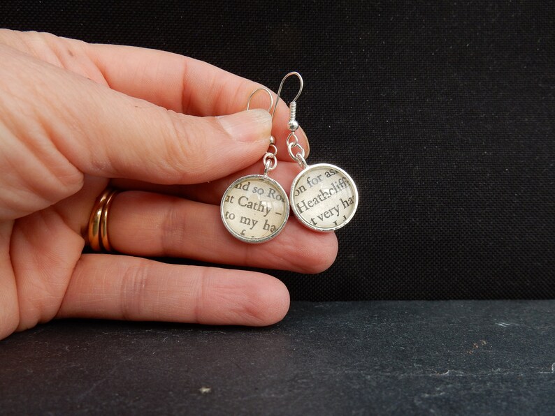 May include: Silver earrings with round glass charms featuring text from the book "Wuthering Heights" by Emily Bront&euml;. The text on the left charm reads "and so Ro at Cathy to my ha", and the text on the right charm reads "on for as Heathcliff t very ha".