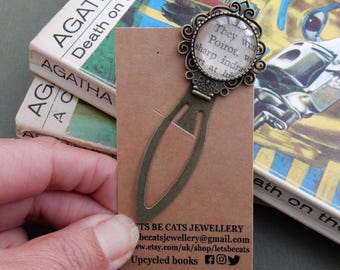 May include: A bronze bookmark with a round, glass charm featuring a quote from Agatha Christie's "Death on the Nile." The bookmark is shaped like a book page and has a decorative design. The charm is surrounded by a decorative metal frame. The bookmark is displayed on a brown card with the text "Lets Be Cats Jewellery" and contact information.