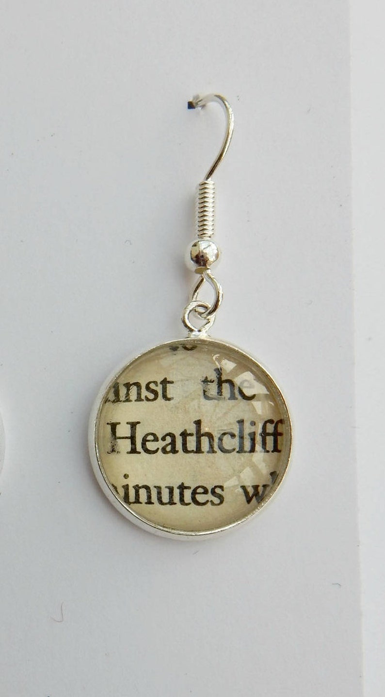 May include: Silver earring with a round glass charm featuring a printed excerpt from the book "Wuthering Heights" that reads "inst the Heathcliff inutes w".