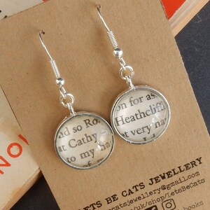 May include: Silver dangle earrings with round glass charms featuring text from the book "Wuthering Heights". The text on the left charm reads "and so Rochester", and the text on the right charm reads "on for as Heathcliff, it very nearly".