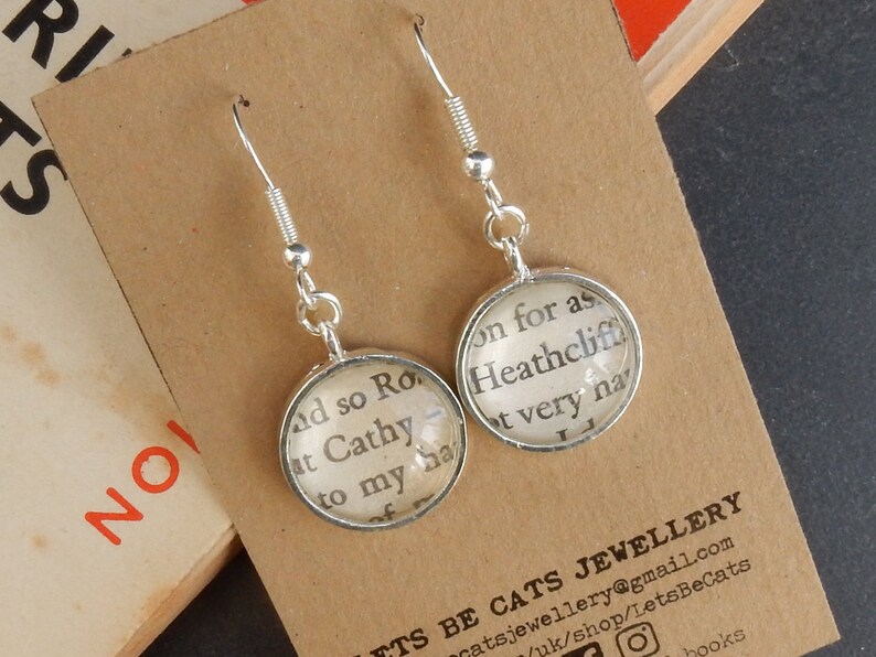 May include: Silver dangle earrings with round glass charms featuring text from the book "Wuthering Heights". The text on the left charm reads "and so Rochester", and the text on the right charm reads "on for as Heathcliff, it very nearly".
