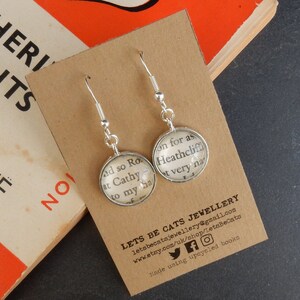 May include: Silver dangle earrings with round glass charms featuring text from the book "Wuthering Heights" by Emily Bront&euml;. The text reads "And so Ro... at Cathy... to my ba..." and "on for as... Heathcliff... t very na..." The earrings are on a brown card with the text "Lets Be Cats Jewellery" and contact information.