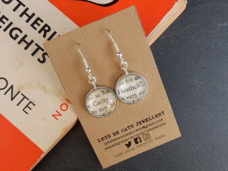 May include: Silver dangle earrings with round glass charms featuring text from the book "Wuthering Heights" by Emily Bront&euml;. The text reads "And so Ro... at Cathy... to my ba..." and "on for as... Heathcliff... t very na..." The earrings are on a brown card with the text "Lets Be Cats Jewellery" and contact information.