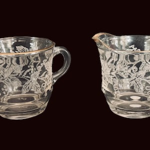 Op de afbeelding: Een suikerpot van helder glas met twee oren en een bijpassend melkkannetje, beide voorzien van een geëtst druivenrankpatroon en een dunne gouden rand langs de bovenkant. De set is gemaakt van transparant glas met een gedetailleerd botanisch ontwerp.