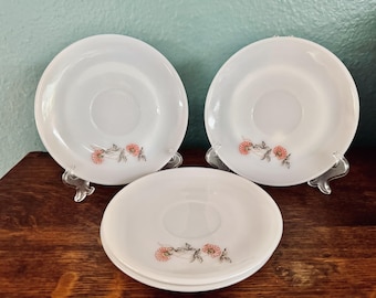 May include: Three white milk glass saucers featuring a delicate floral pattern with small orange-red blossoms and thin gray leaves. Each round saucer has a smooth, glossy finish and a central indentation for a cup, arranged in a group.