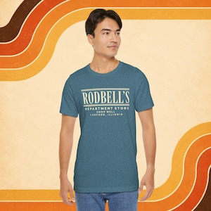 May include: A heathered blue short-sleeve t-shirt featuring the cream-colored text RODBELL'S DEPARTMENT STORE SHOP WELL LANFORD, ILLINOIS printed on the chest in a vintage-inspired font with horizontal lines above and below the name.