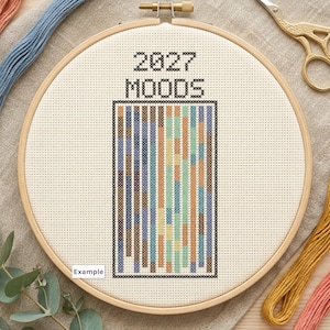 Puede incluir: Un bastidor de bordado circular de madera que contiene un proyecto de punto de cruz sobre tela color crema. El diseño presenta el texto negro 2027 MOODS sobre una cuadrícula rectangular de columnas verticales rellenas de puntos de colores azul, bronce y verde.