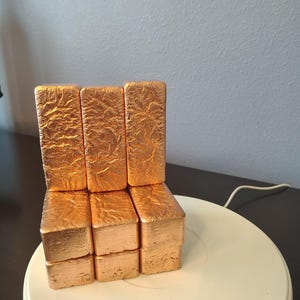 May include: A stack of nine rectangular copper-colored blocks arranged in a three-by-three grid. Each block features a textured, crinkled surface with a metallic sheen, resembling a small chair or seat structure made of individual units.