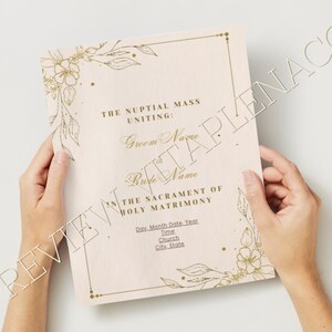 May include: A rectangular wedding program card featuring a pale pink background with gold foil floral illustrations in the corners and a gold border. The text reads: THE NUPTIAL MASS UNITING: Groom Name and Bride Name IN THE SACRAMENT OF HOLY MATRIMONY.