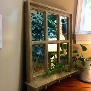 May include: A decorative wooden windowpane mirror with a shelf and hooks. The mirror has multiple panes and reflects a leafy green plant and a lamp.