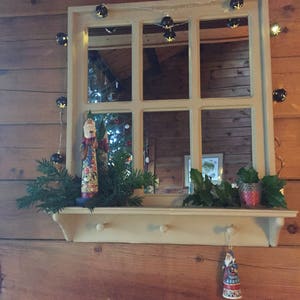 May include: A white wooden shelf with a mirror above it. The mirror has a wood frame and is divided into six panes. There are Christmas lights strung around the mirror and a small Christmas tree in the reflection. There are two small figurines on the shelf, one of Santa Claus and one of a snowman. There are also some green branches on the shelf.