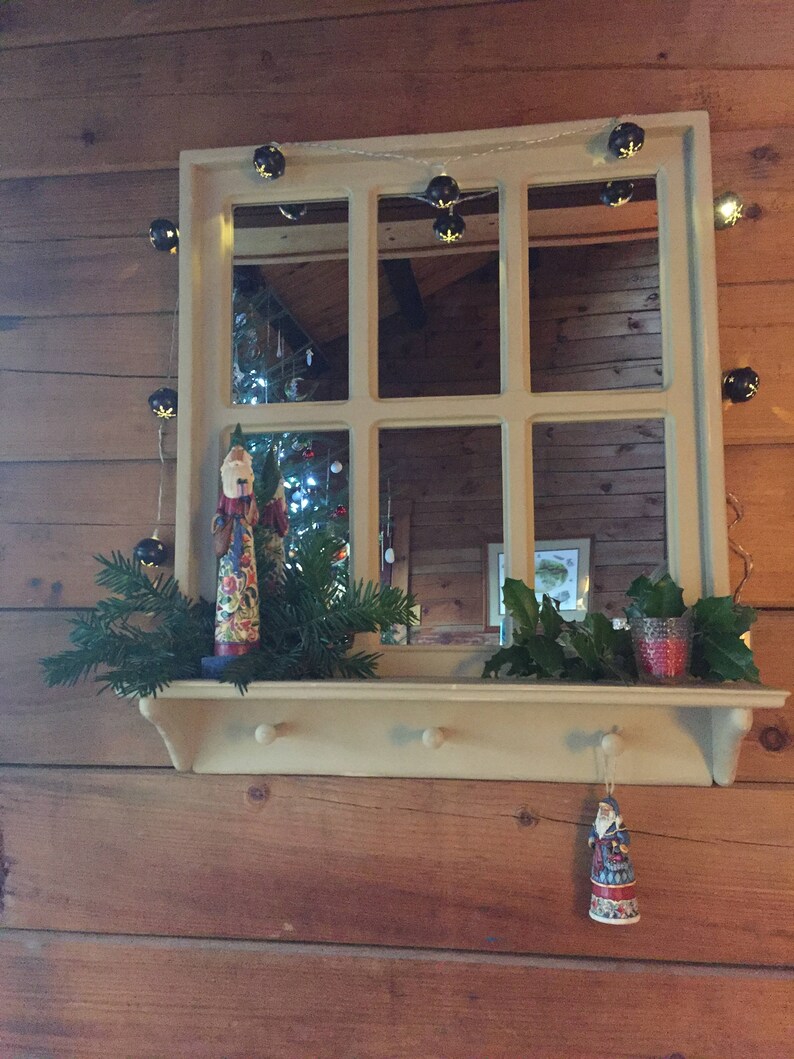 May include: A white wooden shelf with a mirror above it. The mirror has a wood frame and is divided into six panes. There are Christmas lights strung around the mirror and a small Christmas tree in the reflection. There are two small figurines on the shelf, one of Santa Claus and one of a snowman. There are also some green branches on the shelf.