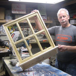 May include: A man holds a wooden window frame with multiple panes of glass. The frame is unfinished and has a natural wood color. The man is wearing a grey t-shirt with the word "KEMP" printed on it in red.