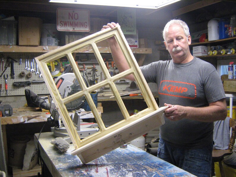 May include: A man holds a wooden window frame with multiple panes of glass. The frame is unfinished and has a natural wood color. The man is wearing a grey t-shirt with the word "KEMP" printed on it in red.