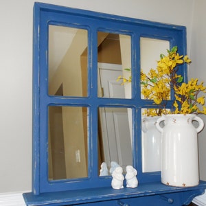 May include: A blue painted wood framed mirror with a shelf and multiple panes. The mirror is reflecting a room with a white door and yellow flowers in a white vase.