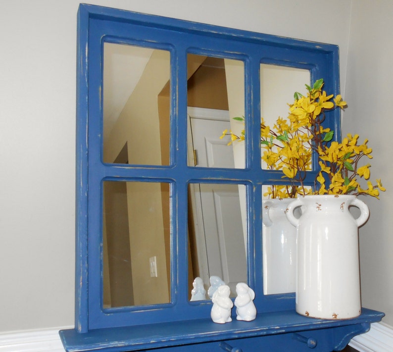 May include: A blue painted wood framed mirror with a shelf and multiple panes. The mirror is reflecting a room with a white door and yellow flowers in a white vase.