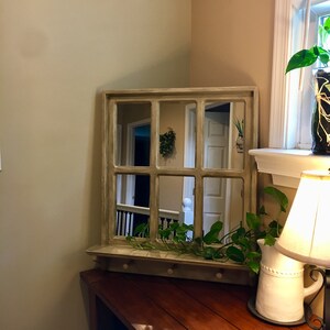 May include: A light brown wooden framed mirror with a shelf and hooks. The mirror is divided into nine panes and reflects a hallway with a white door and a railing. A green plant is hanging from the shelf.
