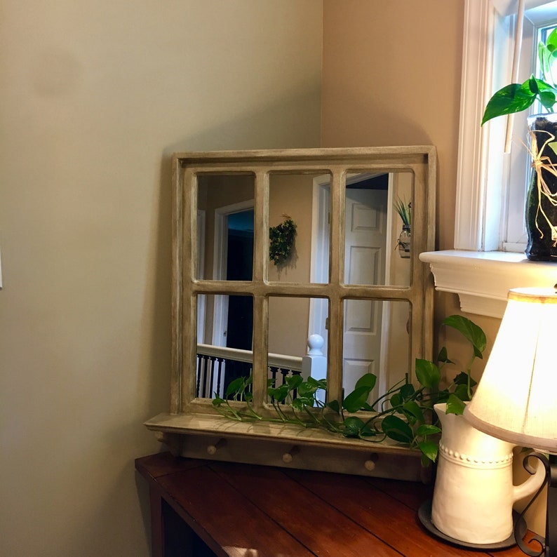 May include: A light brown wooden framed mirror with a shelf and hooks. The mirror is divided into nine panes and reflects a hallway with a white door and a railing. A green plant is hanging from the shelf.