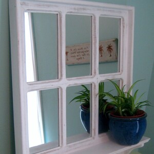 May include: White framed mirror with multiple panes, resembling a window. The mirror is distressed and has a shelf underneath. Two potted plants are on the shelf, one with a blue pot and the other with a green pot. The mirror reflects a wall with a sign that reads "To stay lost is to stay found. Let's been on a margarita voyage."