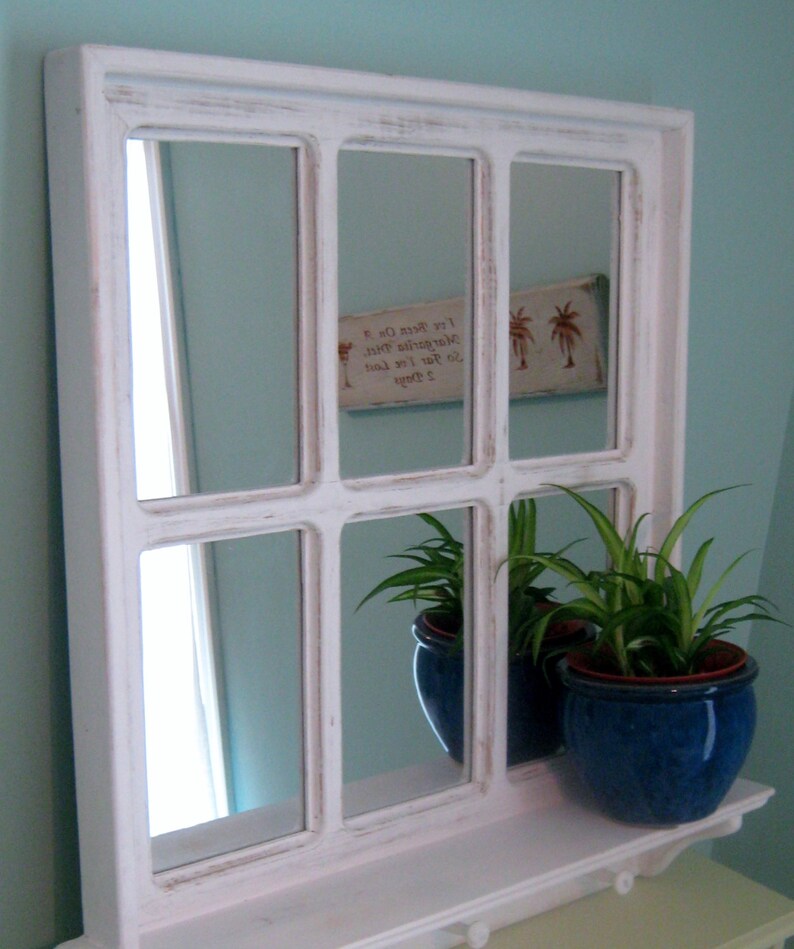 May include: White framed mirror with multiple panes, resembling a window. The mirror is distressed and has a shelf underneath. Two potted plants are on the shelf, one with a blue pot and the other with a green pot. The mirror reflects a wall with a sign that reads "To stay lost is to stay found. Let's been on a margarita voyage."
