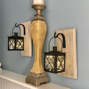May include: Two black metal lanterns with glass panels and internal string lights, each hanging from a black hook mounted on a rectangular light-colored wooden board, positioned on either side of a tall, ornate, beige and gold candle holder.