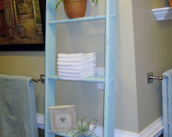 May include: A light blue ladder shelf with three shelves. The top shelf has a green plant in a terracotta pot. The middle shelf has a stack of white towels. The bottom shelf has a small framed picture and a small green plant. The ladder shelf is mounted on a white wall.