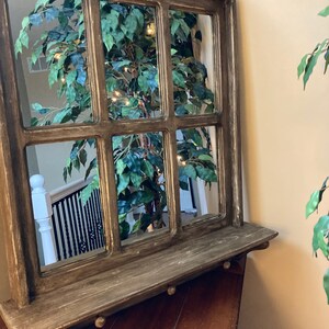 May include: A rustic wooden framed mirror with a shelf. The mirror is divided into nine panes and reflects a green plant with white lights. The frame is painted in a dark brown color with a distressed finish.