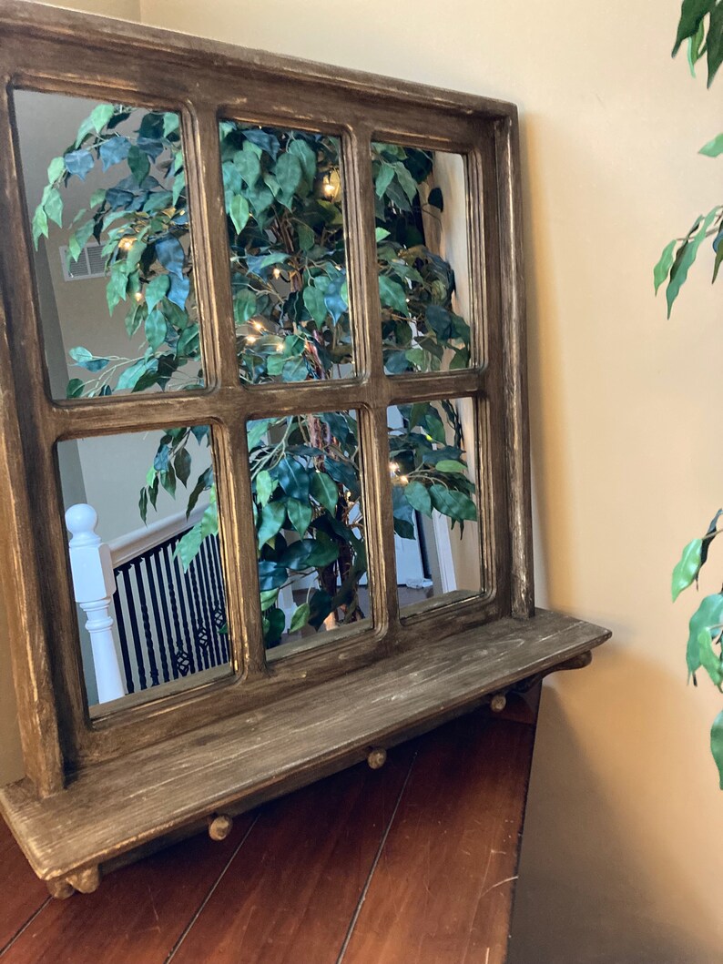 May include: A rustic wooden framed mirror with a shelf. The mirror is divided into nine panes and reflects a green plant with white lights. The frame is painted in a dark brown color with a distressed finish.