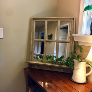May include: A light brown wooden framed mirror with a windowpane design. The mirror is reflecting a hallway with a white door and a green plant. The mirror is mounted on a wooden shelf with hooks. A white pitcher with a beaded design is on the shelf with a green plant.