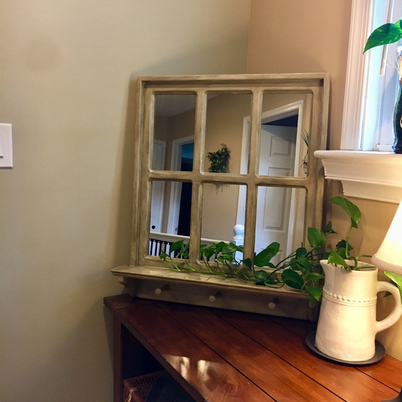 May include: A light brown wooden framed mirror with a windowpane design. The mirror is reflecting a hallway with a white door and a green plant. The mirror is mounted on a wooden shelf with hooks. A white pitcher with a beaded design is on the shelf with a green plant.