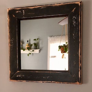 May include: A square wall mirror featuring a thick, distressed black wooden frame with visible light brown wood grain showing through the worn edges. The frame has a wide, flat profile with mitered corners, creating a bold, textured appearance.