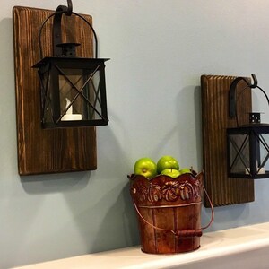 May include: Two black metal lanterns with glass panels are mounted on rectangular dark brown stained wooden boards. A copper-colored metal bucket with a decorative embossed rim sits between the lanterns, filled with several green apples.