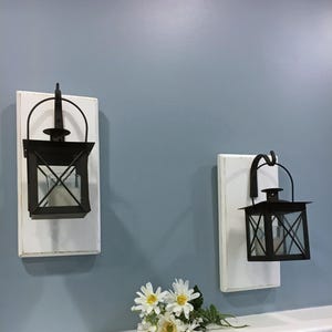 May include: Two wall-mounted sconces featuring rectangular white wooden backboards with distressed edges. Each sconce holds a black metal lantern with a glass insert and a handle, suspended from a black metal hook attached to the wooden board.