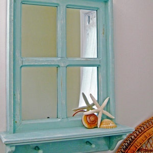May include: A turquoise-painted wooden mirror with a shelf and two hooks. The mirror has a window-like design and is decorated with seashells and starfish. The frame has a distressed finish, giving it a rustic, coastal feel.