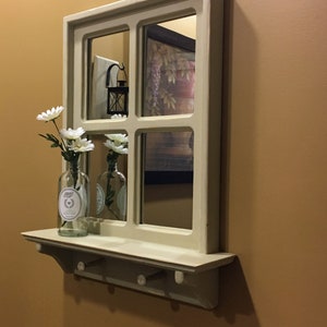 May include: A white wooden windowpane mirror with a shelf and hooks underneath. The mirror has four panes and reflects a room with a painting and a lantern. White flowers are in a clear glass bottle on the shelf.