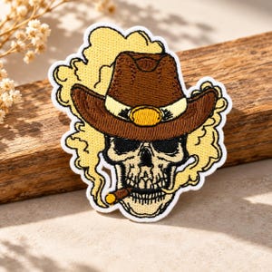Puede incluir: Un parche bordado que muestra una calavera con un sombrero de vaquero marrón con una banda y hebilla amarillas. La calavera tiene un cigarro en la boca y está rodeada por una nube de humo amarillo, todo bordeado con hilo blanco.