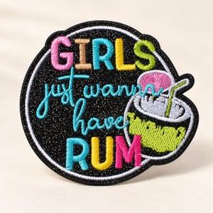 Puede incluir: Un parche bordado circular de color negro brillante con el texto "GIRLS just wanna have RUM" en hilo de colores. La palabra "GIRLS" está en rosa, beige, azul y amarillo, mientras que "RUM" está en azul, amarillo y rosa. Incluye un cóctel verde con una sombrilla rosa.